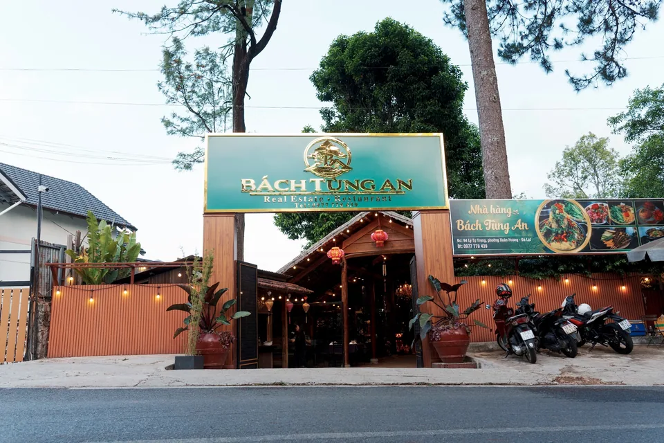Mặt tiền nhà hàng Bò Tơ Bách Tùng An tại 94 Lý Tự Trọng, Đà Lạt — bảng hiệu xanh chữ vàng, lối vào lát gỗ với đèn lồng đỏ và dây đèn vàng giữa tán thông.
